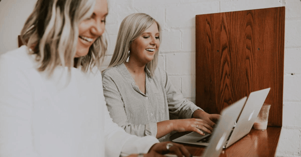 Two women at the office laughing while working on their computers.